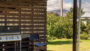 A barbecue grill with a wooden table and chair, set against a wooden fence with a view of a grassy area and trees.