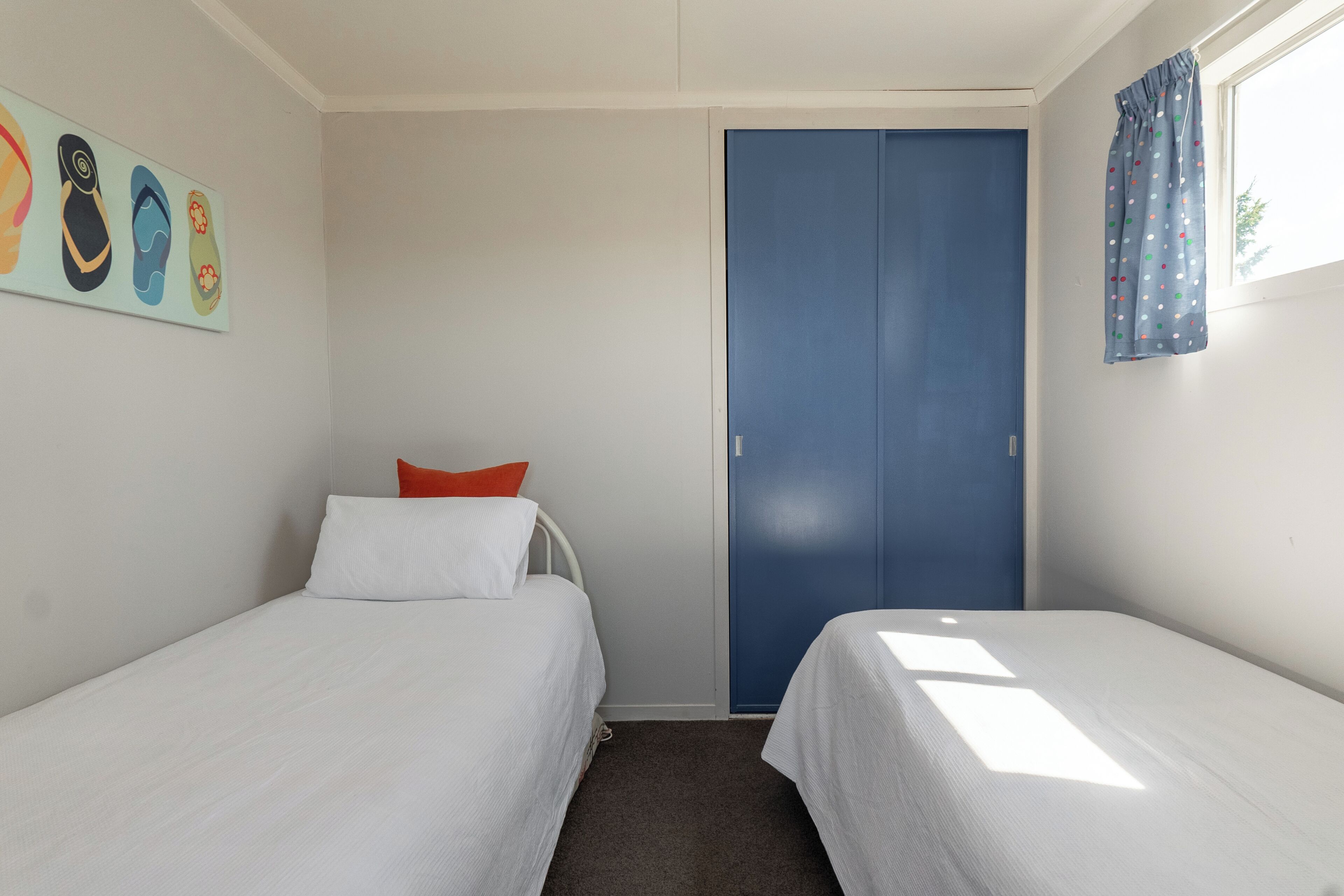 Een kamer met twee eenpersoonsbedden, een blauwe deur, een raam met gordijnen en een muur met kleurrijke kunstwerken.
