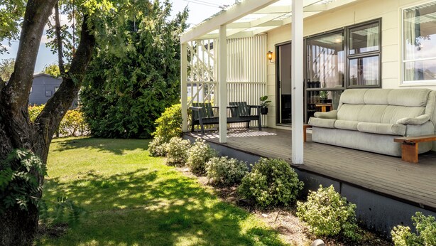 A covered outdoor seating area with a sofa and chairs, a wooden deck, and a garden with grass and bushes. - Classic 3 Bedroom Holiday Home (Twizel)