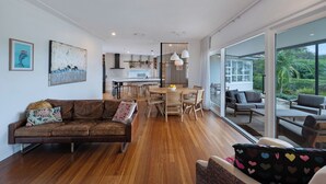 Un interior moderno con suelo de madera, un sofá de cuero marrón, una zona de comedor con mesa redonda y ventanas amplias que dan a zonas verdes.