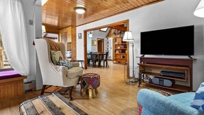 Una sala de estar acogedora con techo de madera, un televisor, un sofá, una silla, un taburete pequeño y un área de comedor al fondo.