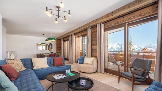 Ein geräumiger Wohnbereich mit einer Couch, einem Sessel und einem runden Tisch. Große Fenster bieten Blick auf schneebedeckte Berge.
