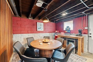 A round wooden table with chairs, a red brick wall, a framed picture, and a small kitchenette with a coffee machine.