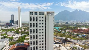 Un edificio alto con varios pisos, rodeado de otros edificios y un paisaje montañoso al fondo.
