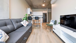 Una sala de estar moderna con un sofá gris, un televisor, un comedor y una cocina al fondo, y un cielorraso con un equipo de aire acondicionado.