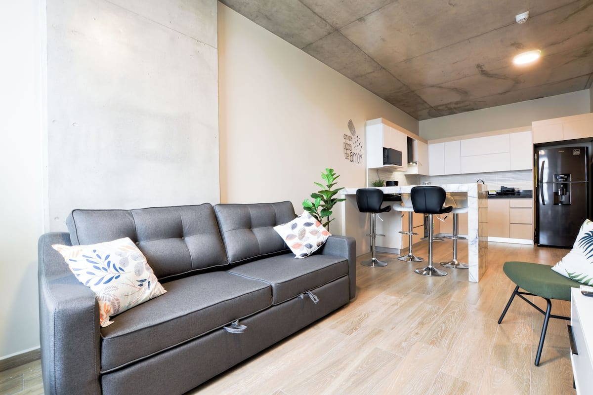 Una sala de estar moderna con un sofá gris, dos taburetes altos, una mesita y una zona de cocina con refrigerador.