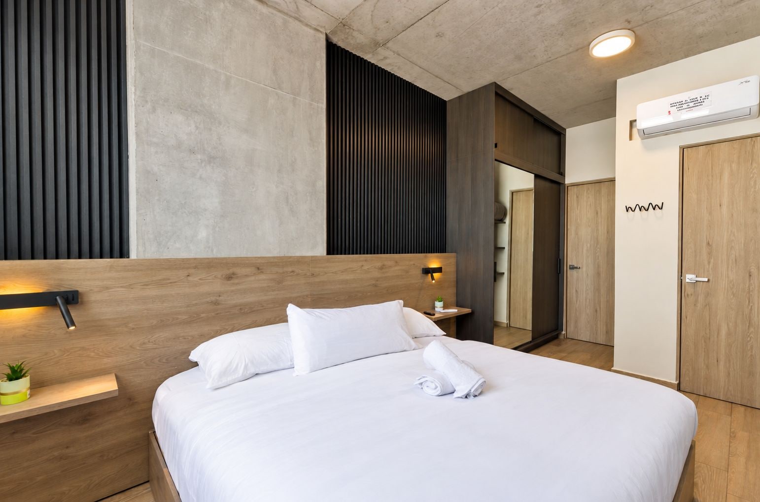 Habitación de hotel moderna con cabecera de madera, mesita de noche con lámpara, cama grande con sábanas blancas, unidad de aire acondicionado montada en la pared y armario.