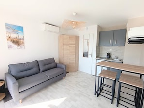 Ein kompakter Wohnraum mit grauem Sofa, einer kleinen Küchenzeile und einem Esstisch mit Hocker.