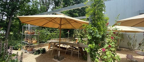 An outdoor patio area with a table and chairs under a large umbrella, surrounded by greenery and a gravel ground.