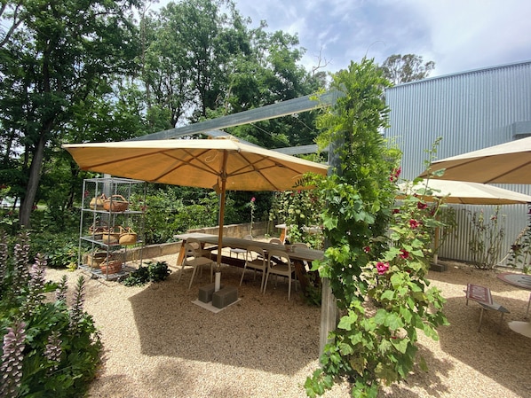 An outdoor patio area with a table and chairs under a large umbrella, surrounded by greenery and a gravel ground.