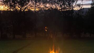 A fire pit with flames, surrounded by a concrete surface, with a grassy area and trees in the background.