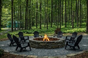 Een ronde vuurkuip met vuur, omringd door zes Adirondack-stoelen, gelegen op een grindpad in een bos.