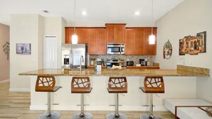 A modern kitchen with wooden cabinets, a central island with bar stools, stainless steel appliances, and pendant lighting. - Private Pool 4BR Getaway in ChampionsGate (Four corners)