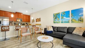 A modern living room with a sofa, chairs, a coffee table, and a kitchen area with wooden cabinets and a refrigerator. - Private Pool 4BR Getaway in ChampionsGate (Four corners)