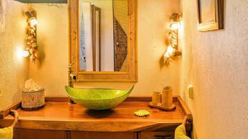 A bathroom with a wooden vanity, a green glass bowl sink, a mirror, and wall-mounted lights.