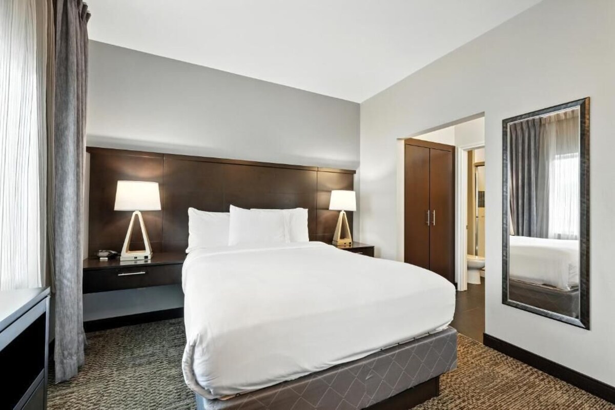 Una habitación de hotel con una cama grande, dos mesitas de noche con lámparas, un cabecero de madera oscura, un espejo en la pared y una puerta que da al baño.