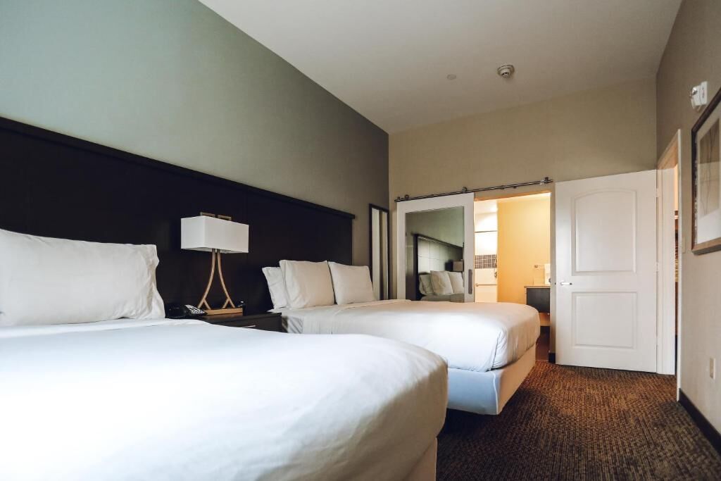 A hotel room with two beds, a nightstand with a lamp, and a door leading to a bathroom.