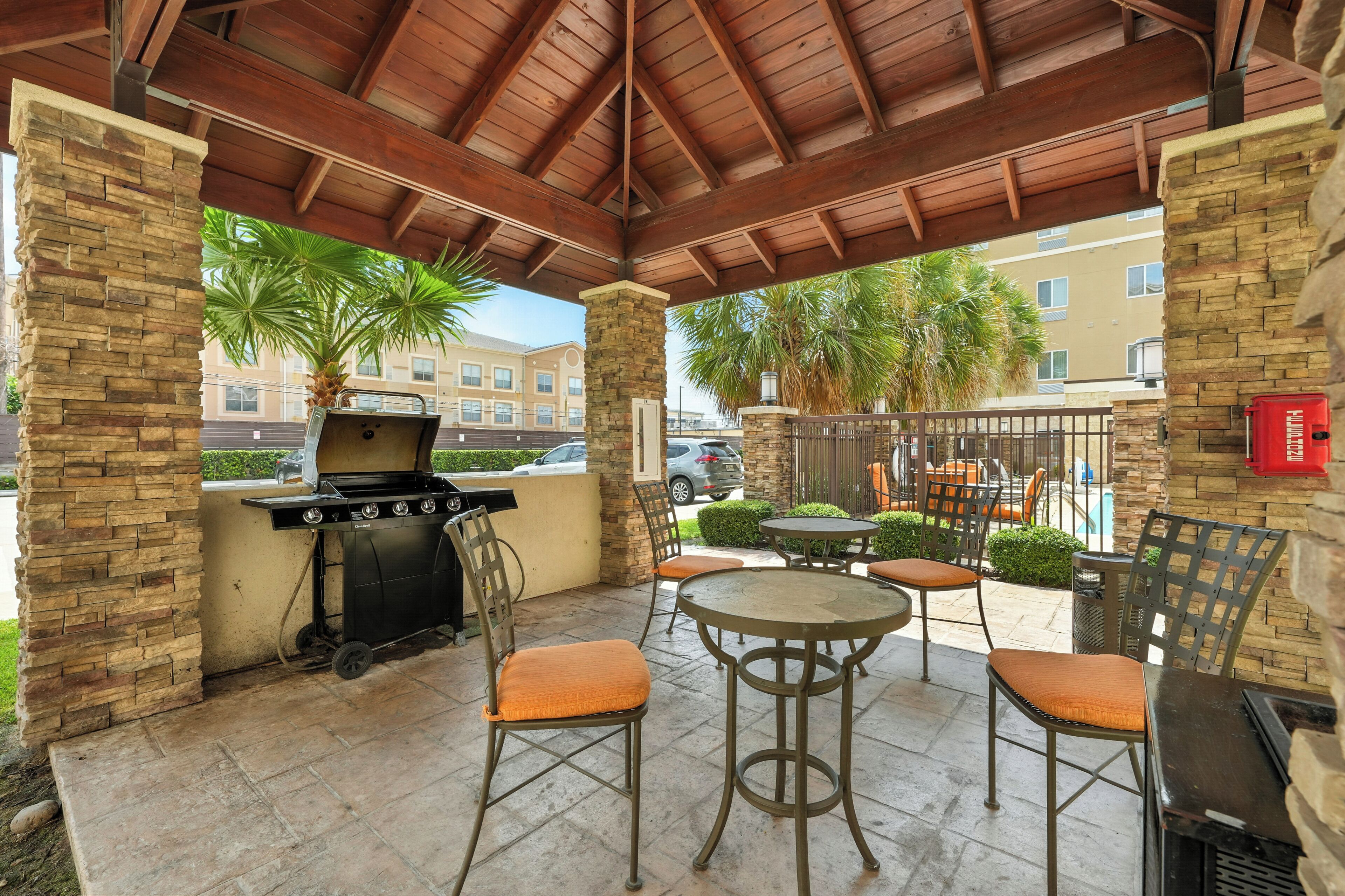 An outdoor patio with a grill, chairs, and tables, surrounded by stone walls and palm trees.