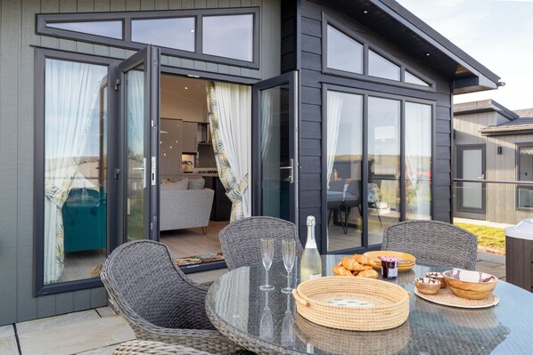 Un espace de repas extérieur moderne, avec des chaises en osier, une table en verre et un jacuzzi en arrière-plan.