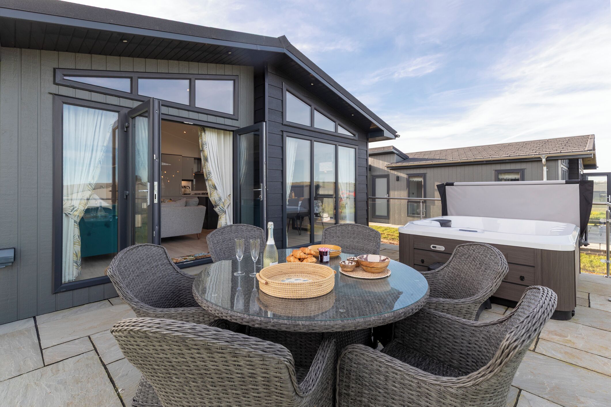A modern outdoor dining area with wicker chairs, a glass table, and a hot tub.