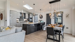 A modern kitchen with a central island, dining area with a table set for four, and a living area with a sofa. - Bryn Hafod - Luxury Lodge, Hot Tub, Walk to Beach (Penally)