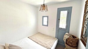 Une petite chambre avec un lit simple, un mur blanc uni, une porte avec une fenêtre et un luminaire suspendu.