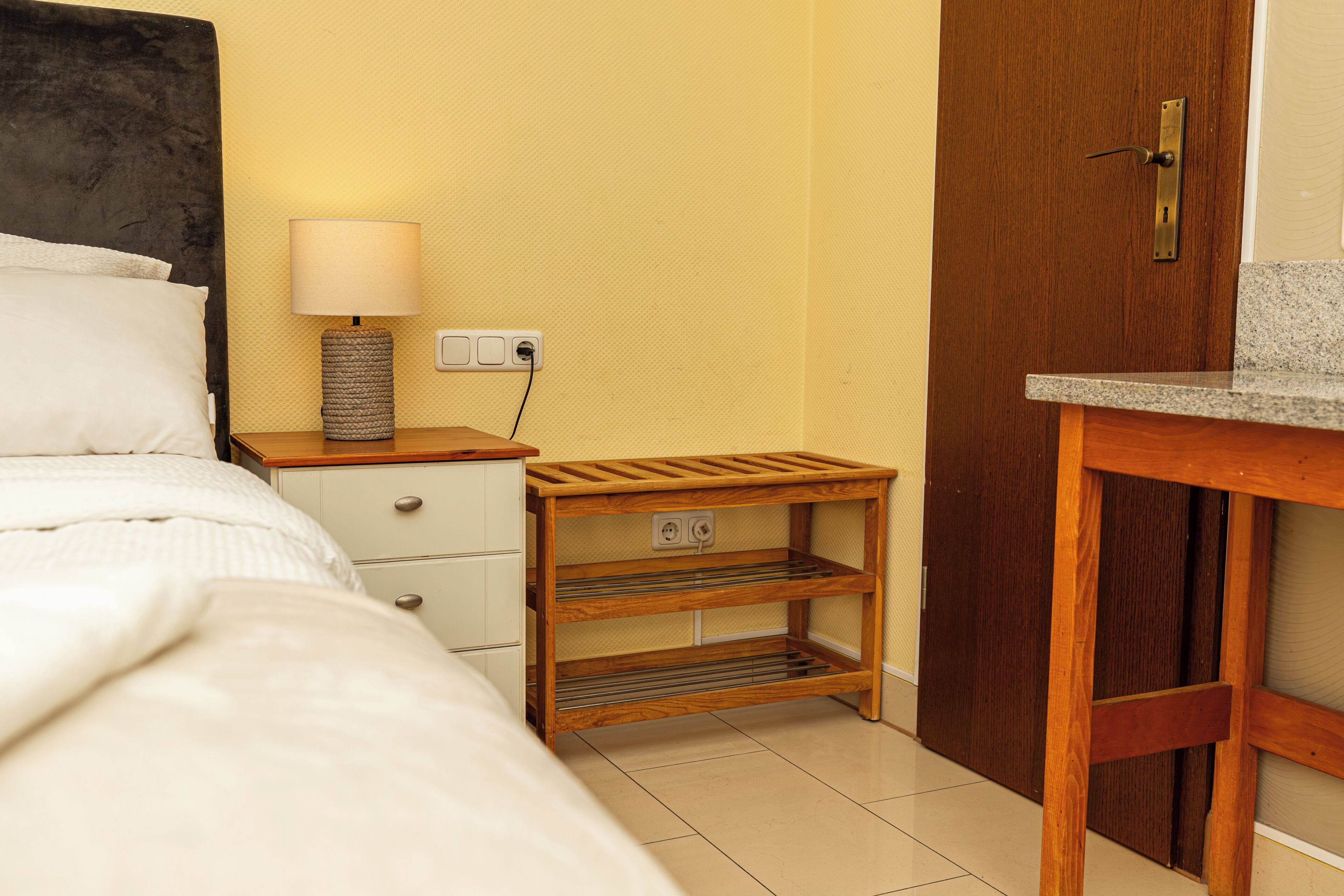 Ein Hotelzimmer mit Bett, Nachttischlampe, einem Holznachttisch und einem kleinen Holzregal.