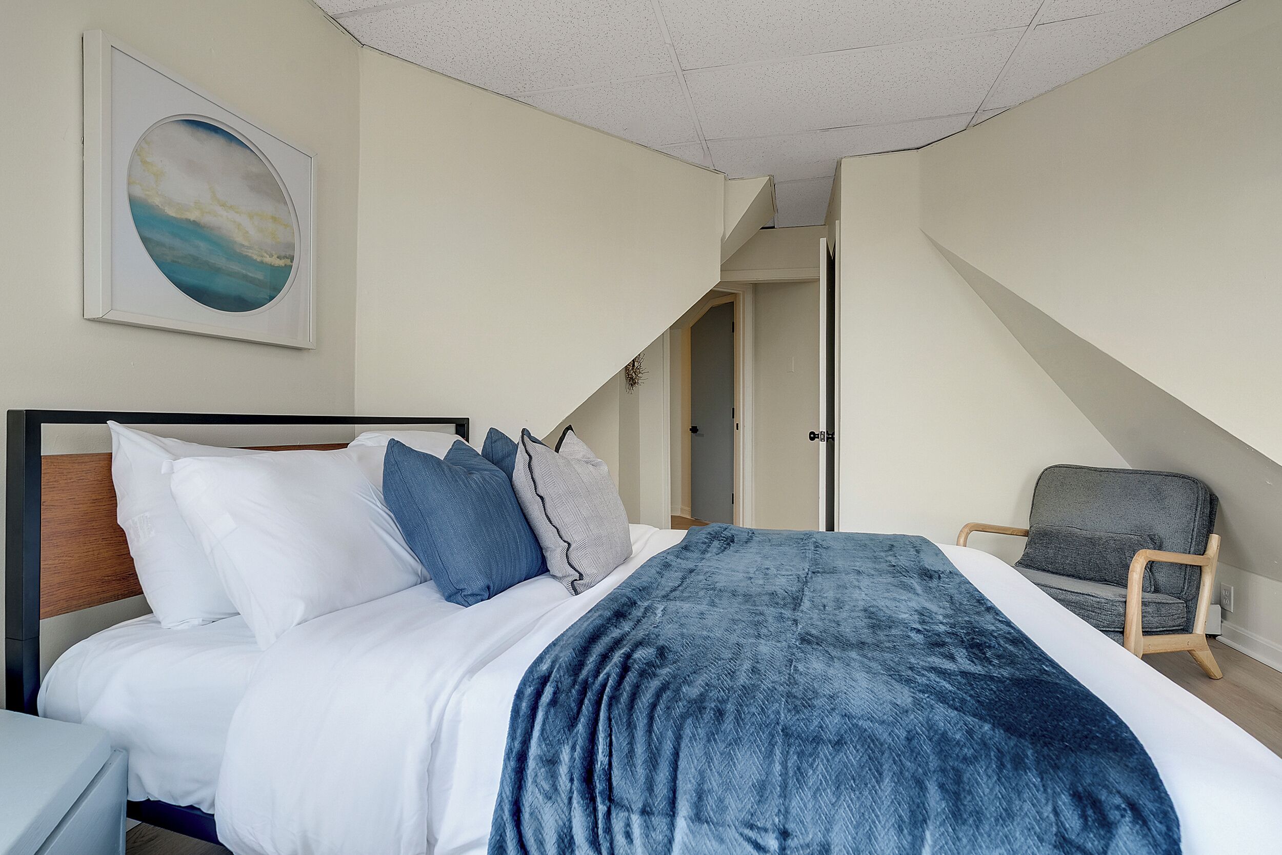 A bedroom with a bed, pillows, a blanket, a chair, and a painting on the wall.