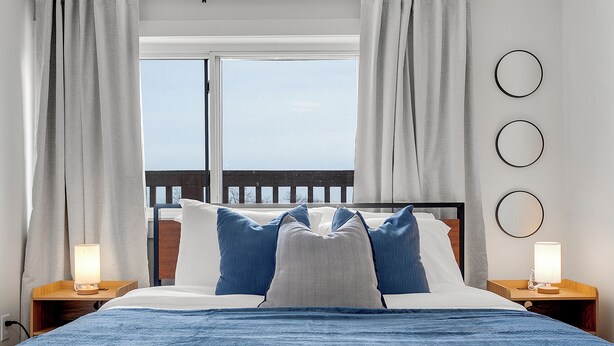 Una cama con una manta azul, dos lámparas de noche y vista al mar a través de una ventana.