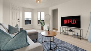 Una sala de estar con un sofá, una mesa de centro, un televisor sobre un mueble y vistas al exterior.