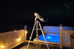Un telescopio sobre un trípode en una terraza de madera, con un jacuzzi al fondo.