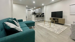 Een moderne woonkamer met een smaragdgroene bank, een flatscreen televisie en een keuken op de achtergrond.
