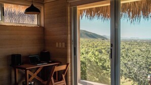 A bedroom with a bed, a desk, and a chair, a large window with a view of a landscape, and a thatched roof. - Hotel Cabanas uno (El Pescadero)