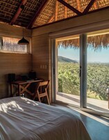 Un dormitorio con cama, escritorio y silla, un ventanal con vistas al paisaje, y un techo de paja.