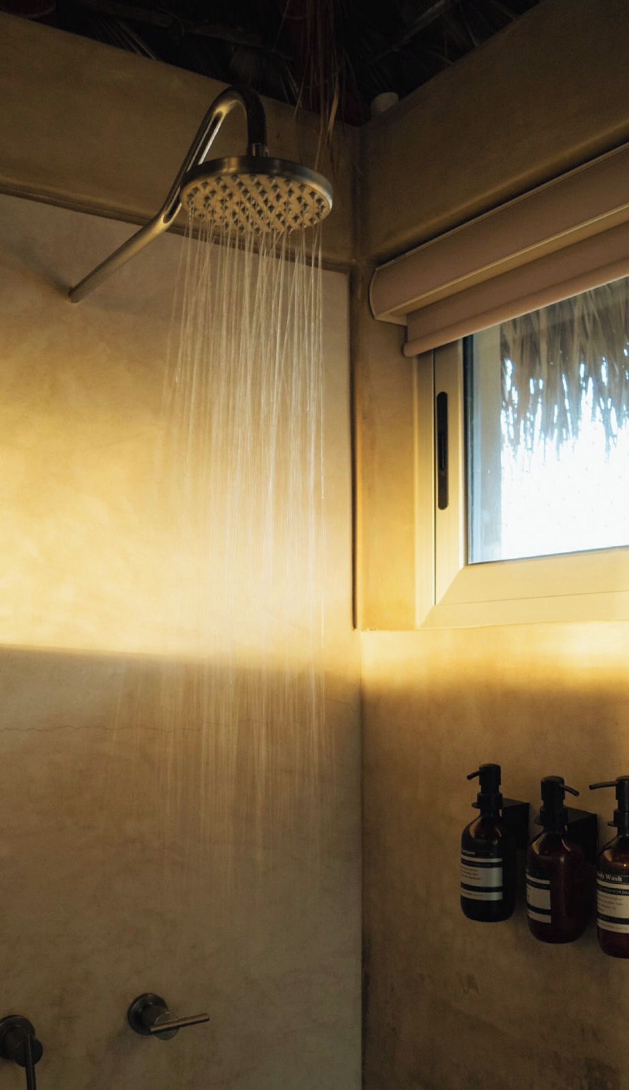 A shower with water running, a window with blinds, and three bottles of soap on the wall.