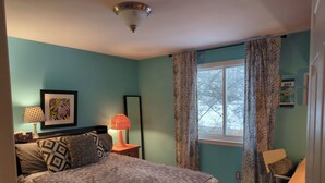Un dormitorio con cama, mesita de noche, lámpara, un cuadro enmarco y una ventana con cortinas.