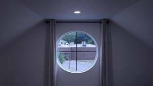 A room with a large circular window, a ceiling light, and a view of a building with trees and a parking lot.
