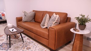 A brown leather sofa with patterned cushions, a small wooden side table with a white vase, a potted plant, and a floral-patterned rug.