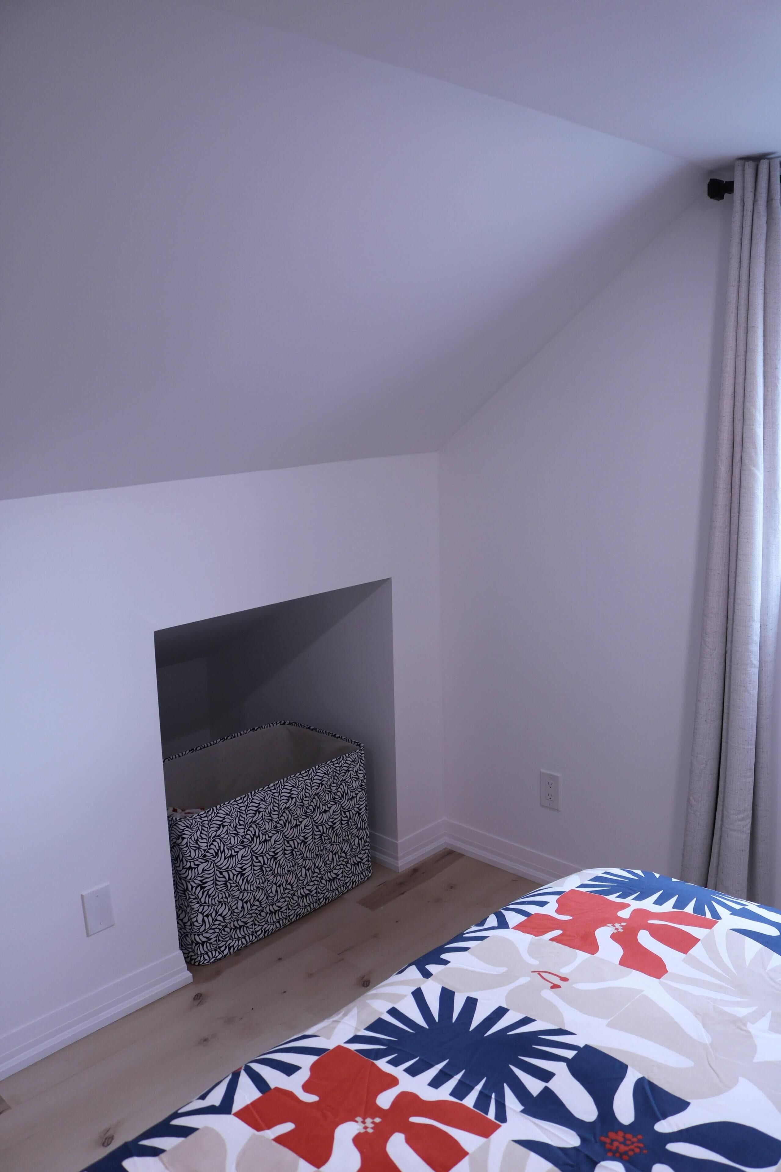 Ett sovrum med ett sluttande tak, en säng med ett färgglatt, blommigt överkast, en mönstrad förvaringslåda och ett fönster med gardiner.