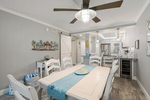 Ein Essbereich mit einem Tisch für zwei Personen, weißen Stühlen, einem Deckenventilator und einer Küche im Hintergrund.