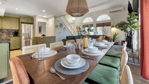 Un comedor con una larga mesa de madera dispuesta para cuatro, rodeada de sillas acolchadas, una lámpara colgante y vistas de la cocina y el área de estar.