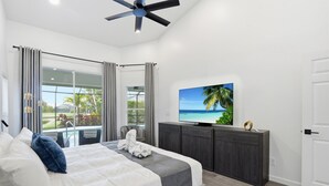 A bedroom with a bed, a TV, a ceiling fan, and a view of a pool and greenery. - 2026 Renovated Luxury Home. Heated Pool. 10 mins to 5th Ave, Pier Beach (Naples)