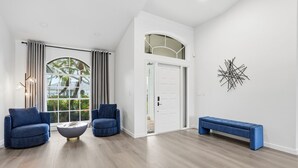 A modern living room with two blue armchairs, a round coffee table, a blue bench, a white door, and a large window with curtains. - 2026 Renovated Luxury Home. Heated Pool. 10 mins to 5th Ave, Pier Beach (Naples)