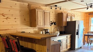 A wooden interior with a kitchen area featuring a central island, bar stools, and a refrigerator. - Beautiful 3-bedroom barn apartment right next to Yellowstone National Park (Gardiner)
