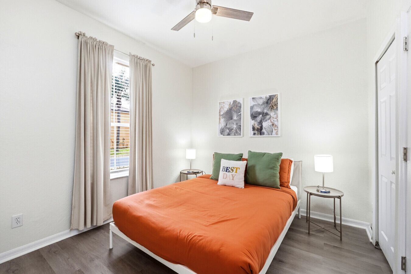 Een slaapkamer met een bed, twee nachtkastjes, een plafondventilator, een raam met gordijnen en twee aan de muur hangende kunstwerken.