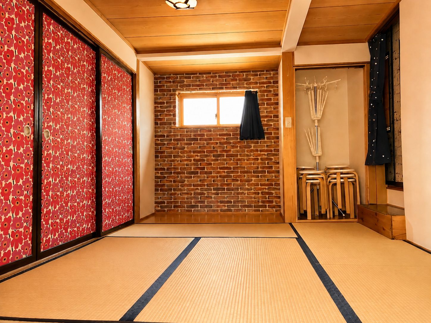 Een kamer met tatami matten, een bakstenen muur, schuifdeuren met rode bloemenmotieven en een klein raam met een zwart gordijn.