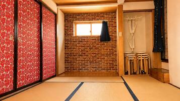 Een kamer met tatami matten, een bakstenen muur, schuifdeuren met rode bloemenmotieven en een klein raam met een zwart gordijn.