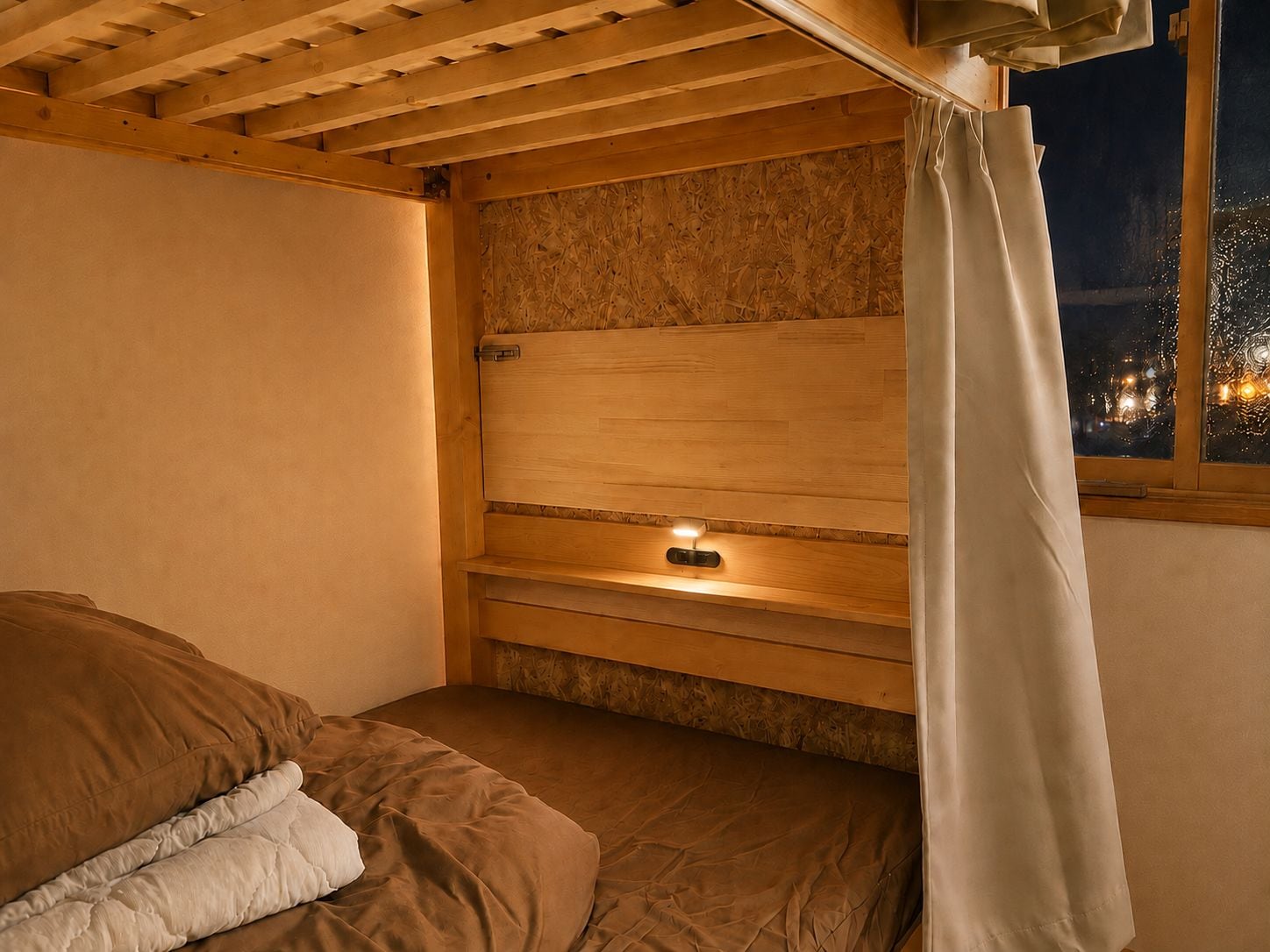 A single bed with a wooden headboard, a bedside table with a lamp, and a window with a view of the city at night.