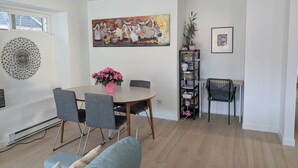 Un coin repas avec une table ronde, des chaises grises, un vase avec des fleurs roses, un tableau accroché au mur, une bibliothèque et une chaise noire.