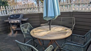 Une table ronde en bois, entourée de chaises en osier, un parasol et un barbecue en arrière-plan.